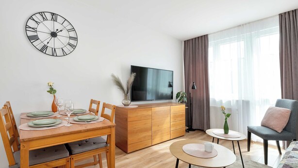 Un saló modern amb una taula de dinar de fusta, cadires, un televisor, un sofà i un rellotge de paret gran.