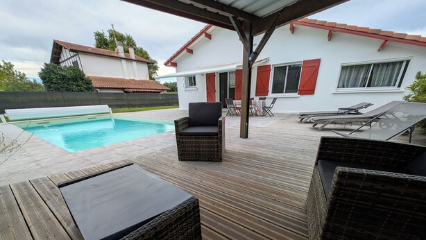 Eine Holzterrasse mit Gartenmöbeln, einem Schwimmbad und einem weißen Gebäude mit roten Fensterläden.