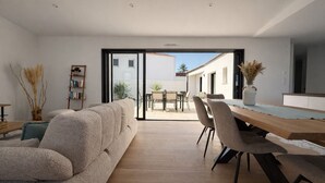 Uma sala de estar moderna com sofá, mesa de jantar, cadeiras e vista para um pátio externo através de uma porta de vidro.