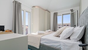 Een slaapkamer met een bed, nachtkastje, gordijnen en uitzicht op gebouwen.