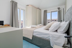 A bedroom with a bed, bedside table, curtains, and a view of buildings outside. - Haller Suite Apartment with Sea View, Wi-Fi, and Air Conditioning (Golfo Aranci)