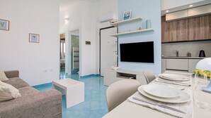 Een moderne woonkamer met een bank, een eettafel gedekt voor twee personen, een flatscreen televisie aan de muur en een keuken met houten keukenkastjes.