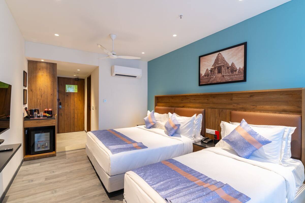 A hotel room with two beds, a wooden headboard, a ceiling fan, a wall-mounted air conditioner, a small kitchenette, and a framed picture of a temple.