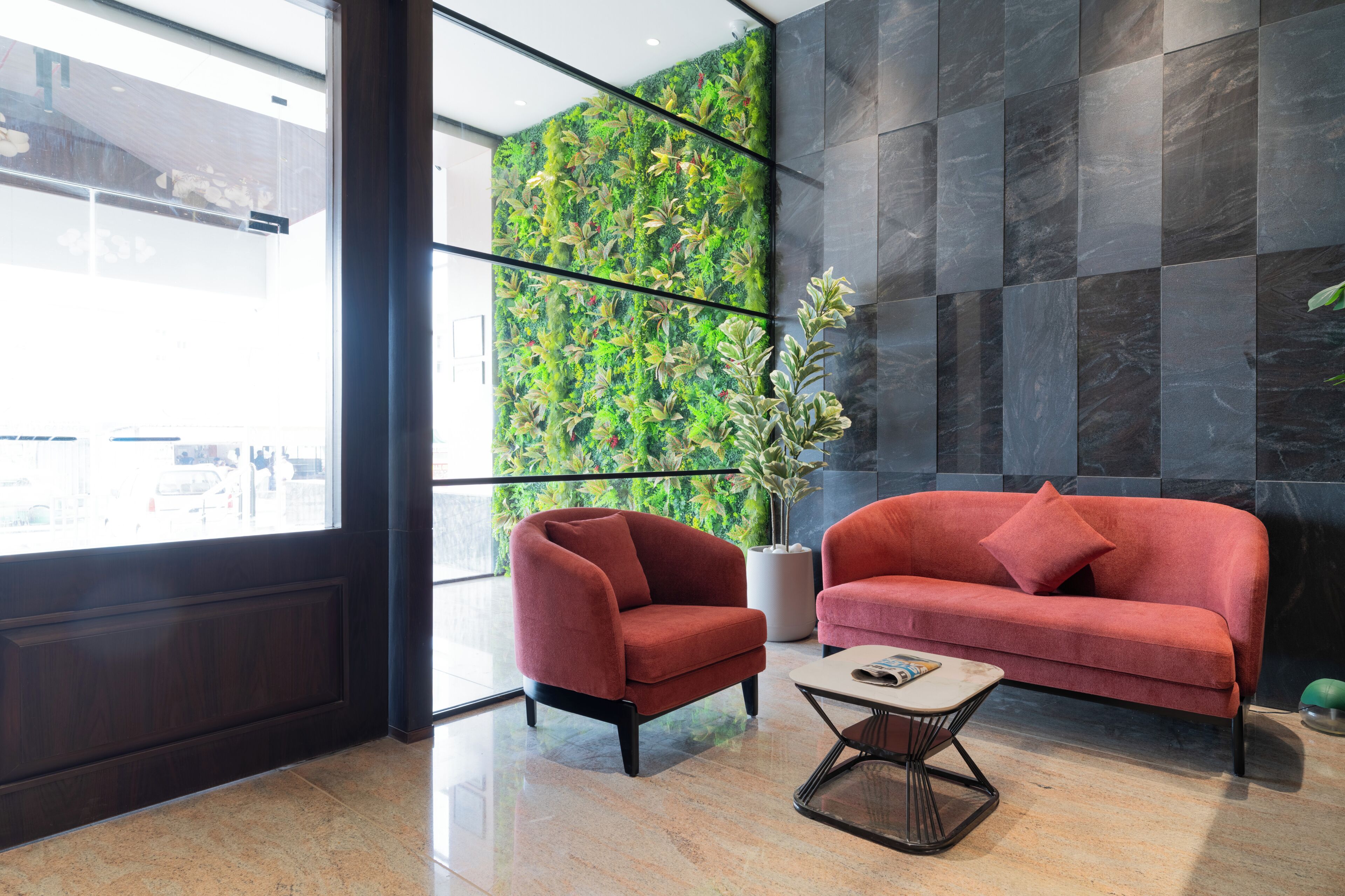 Un vestíbulo moderno con un sofá rojo, un sillón rojo, una mesita de centro, un ventanal grande y una pared verde con plantas.