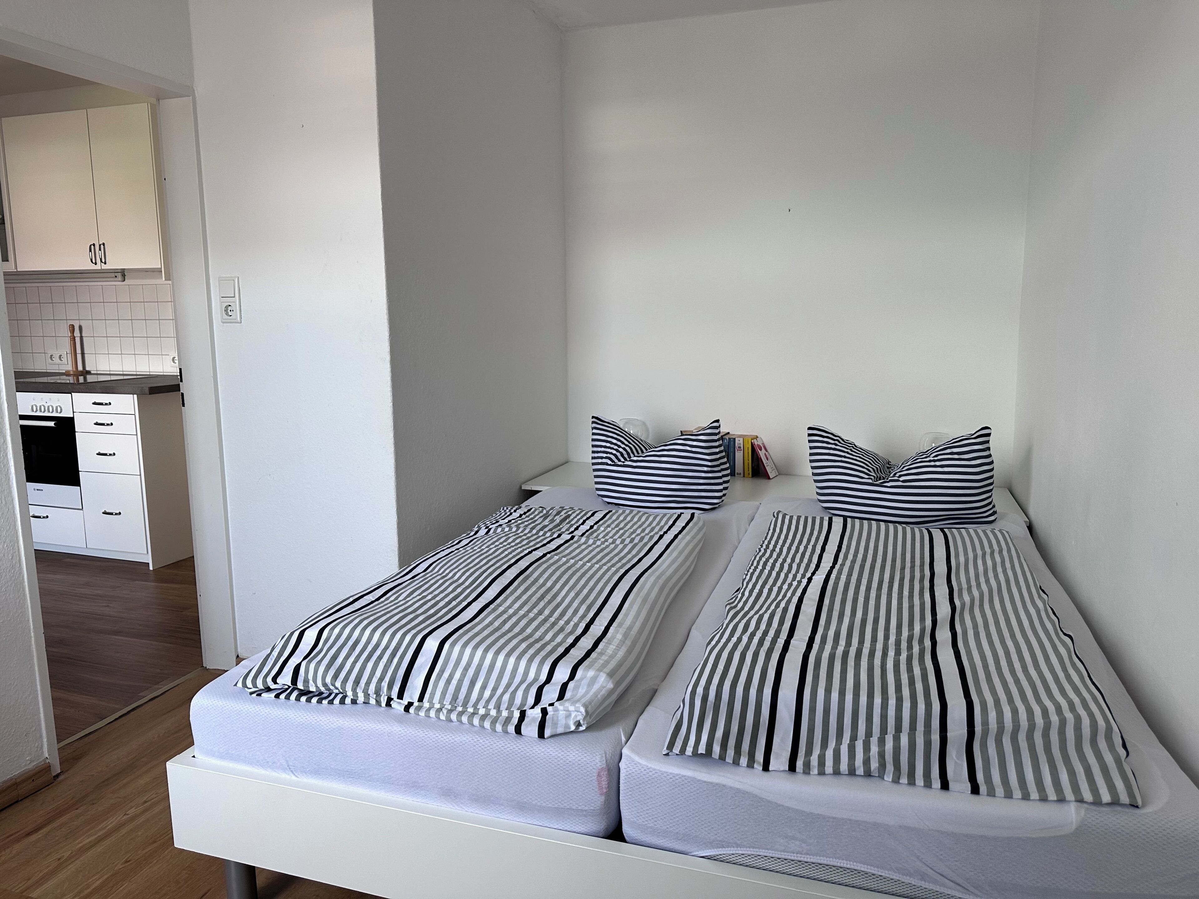 Una habitación pequeña con dos camas individuales, un rincón de cocina y una pared blanca.