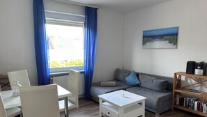 Un salon avec un canapé gris, une table basse blanche, des chaises blanches, une bibliothèque et une fenêtre avec des rideaux bleus.