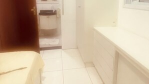 A narrow hallway with a bed on the left, a bathroom with a mirror and sink on the right, and a window with blinds. - Comfort and fun. Beachfront. Small pets are accepted. (Itanhaém)