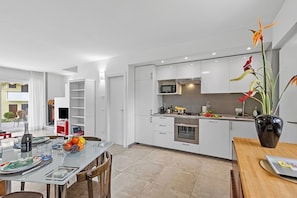 A modern kitchen with white cabinetry, a dining area with a glass table, and a vase with flowers. - Olimpia Home - Terrace Dining and Pool (Tenno)