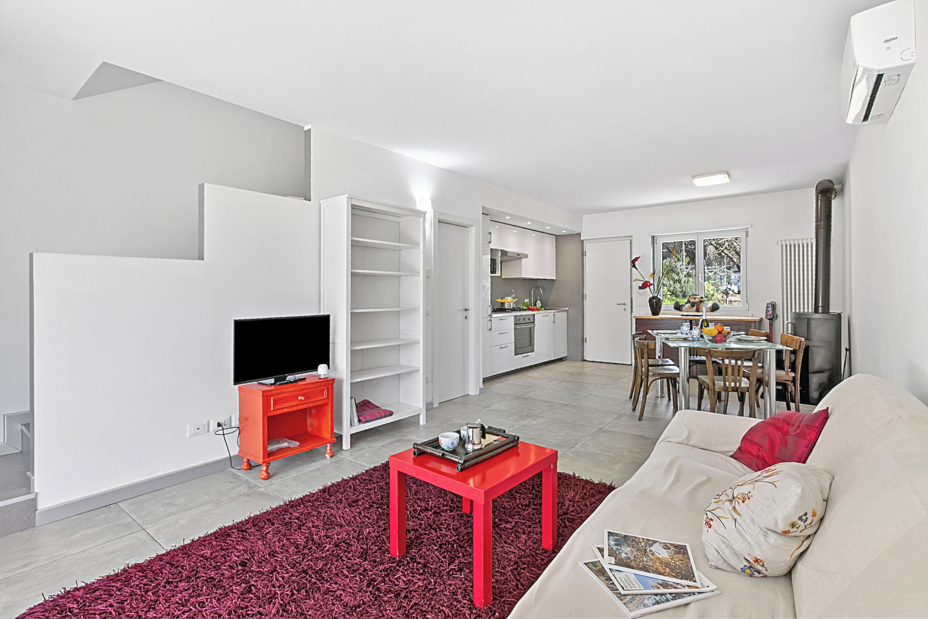 A modern living room with a TV, red coffee table, and sofa, connected to a kitchen with white cabinets and a dining area.