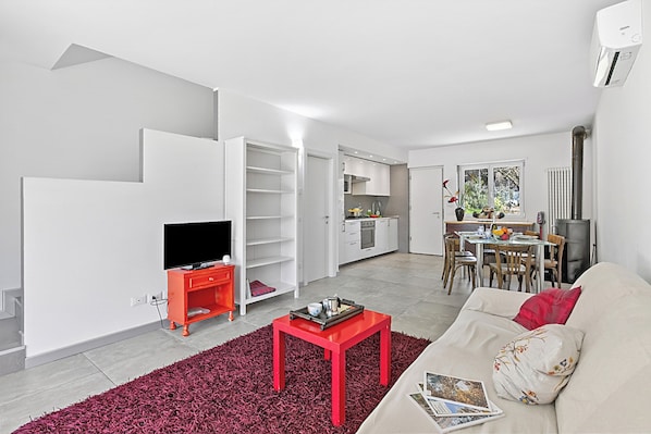 A modern living room with a TV, red coffee table, and sofa, connected to a kitchen with white cabinets and a dining area. - Olimpia Home - Terrace Dining and Pool (Tenno)