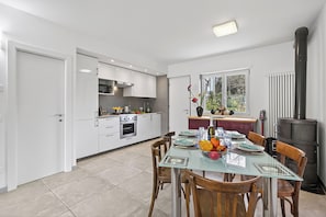A modern kitchen with white cabinets, a dining table with chairs, and a wood-burning stove. - Olimpia Home - Terrace Dining and Pool (Tenno)