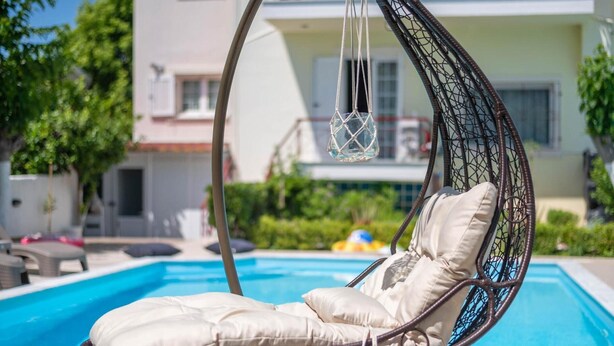 Una silla colgante junto a una piscina con un cojín, una lámpara colgante y un edificio al fondo.