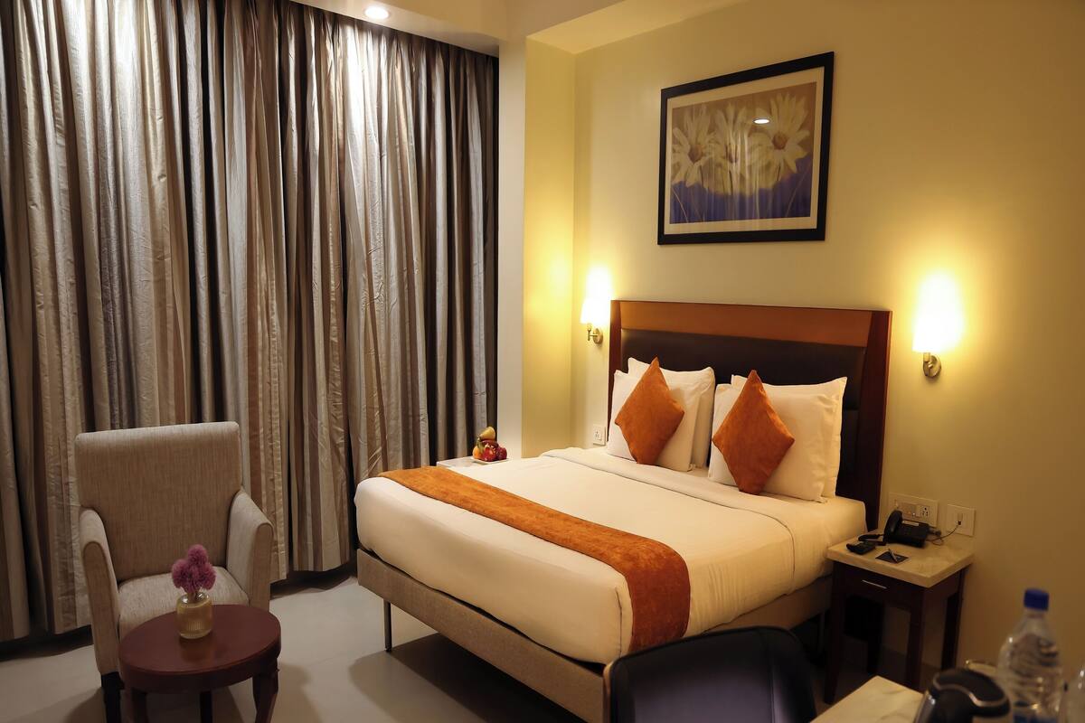 A hotel room with a bed, a chair, a small table with a vase, a nightstand with a phone, and a wall-mounted picture.