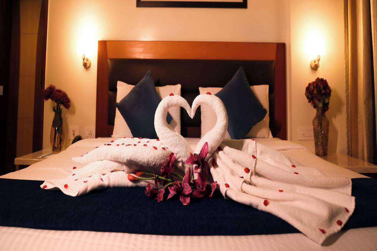 A hotel room with a bed, pillows, a framed floral painting, and a vase with flowers.