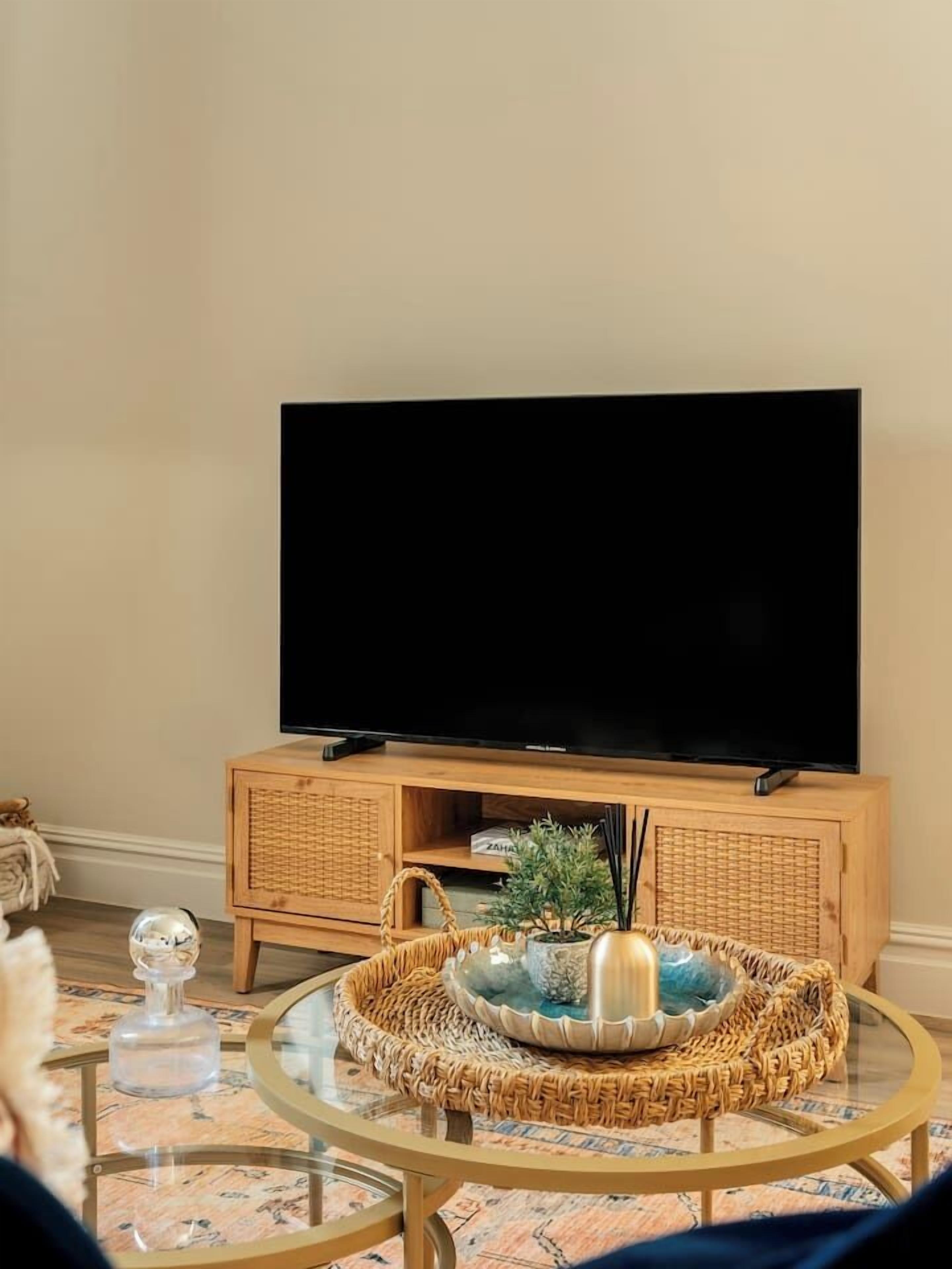 A living room with a flat-screen TV on a wooden cabinet, a glass coffee table with decorative items, and a patterned rug.