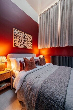 Une chambre à coucher comprenant un lit, des oreillers, une table de chevet, une lampe, une œuvre d’art accrochée au mur et une fenêtre avec des rideaux.