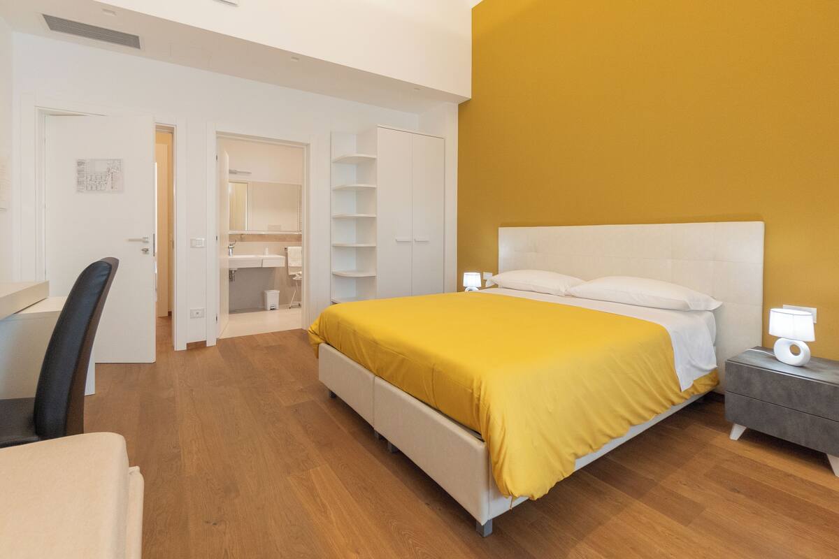 A bedroom with a bed, bedside table, chair, wardrobe, and a bathroom visible through an open door.