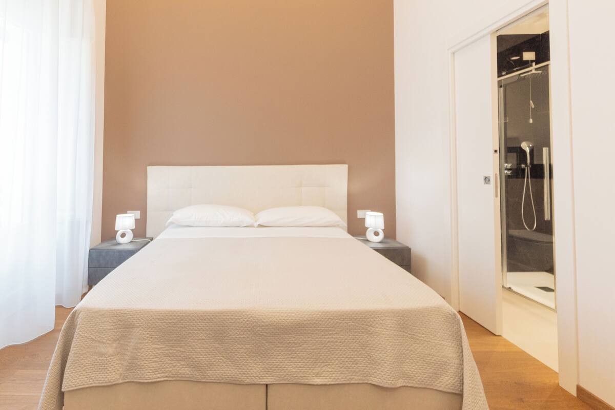 A neatly made bed with a headboard, bedside tables with lamps, and a glass-enclosed shower area.