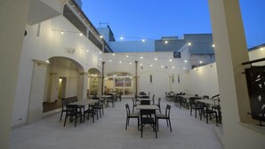 Un espacio al aire libre para comer con mesas y sillas, luces colgantes y vistas a edificios en el fondo.