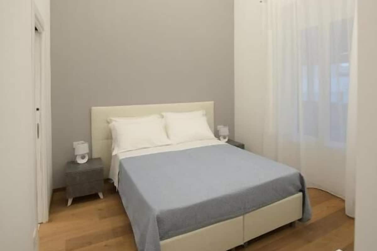 A bedroom with a bed, bedside tables, a nightstand, a ceiling light, and a window with curtains.