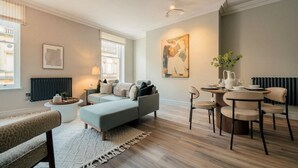A modern living room with a sofa, a dining table with chairs, a radiator, and a window with curtains. - Interior Designed Town Centre One Bed Apartment (North Yorkshire)