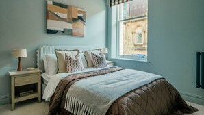 A bedroom with a bed, two bedside tables, a window with a view, and a wall-mounted artwork. - Interior Designed Town Centre One Bed Apartment (North Yorkshire)