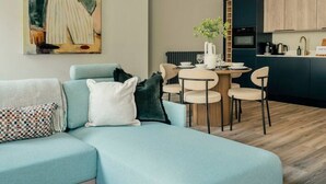 A modern living room with a sectional sofa, a dining area with a round table, and a kitchen with dark cabinetry. - Interior Designed Town Centre One Bed Apartment (North Yorkshire)