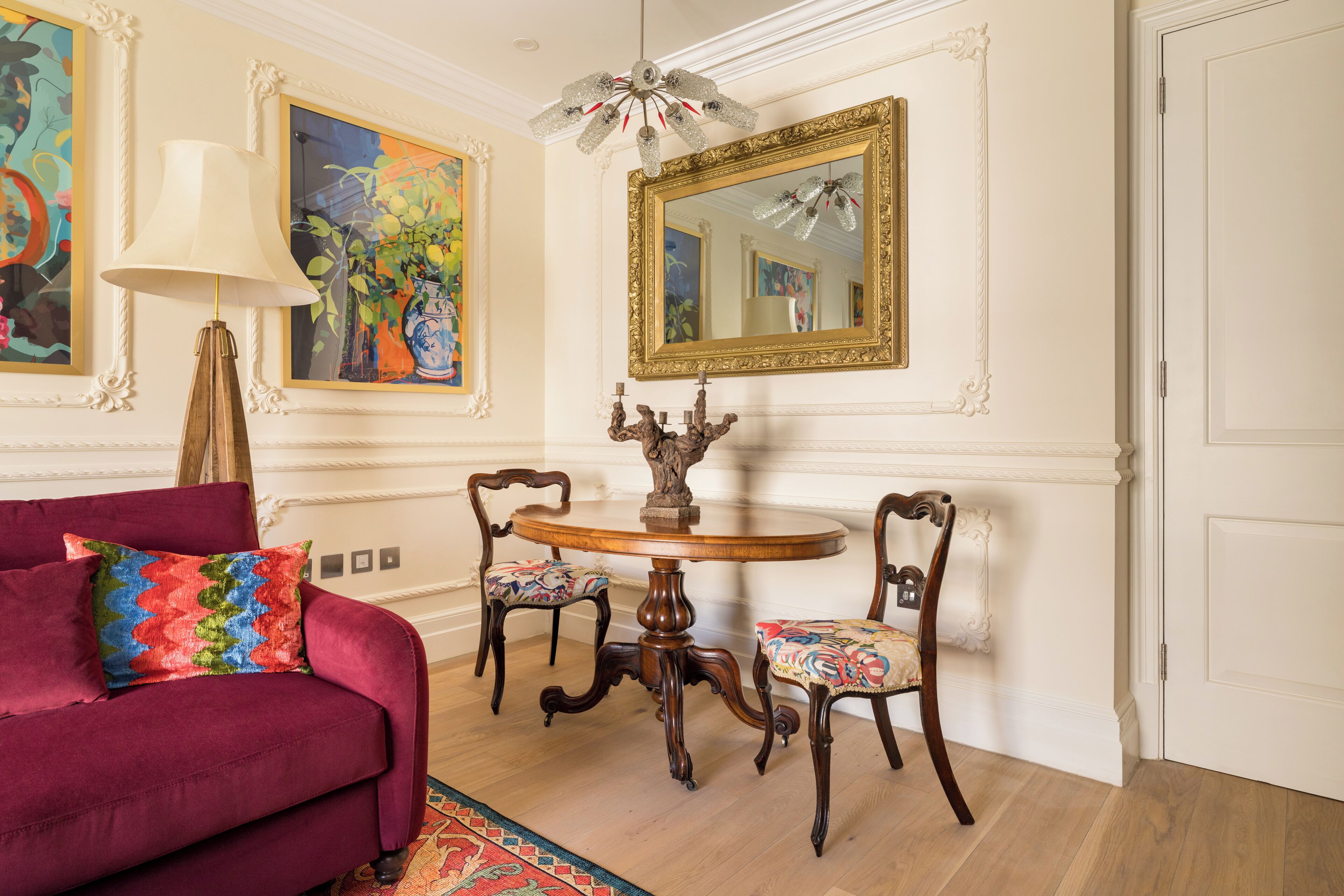 A room with a round wooden table, two chairs, a red sofa, a lamp, and a large mirror on the wall.