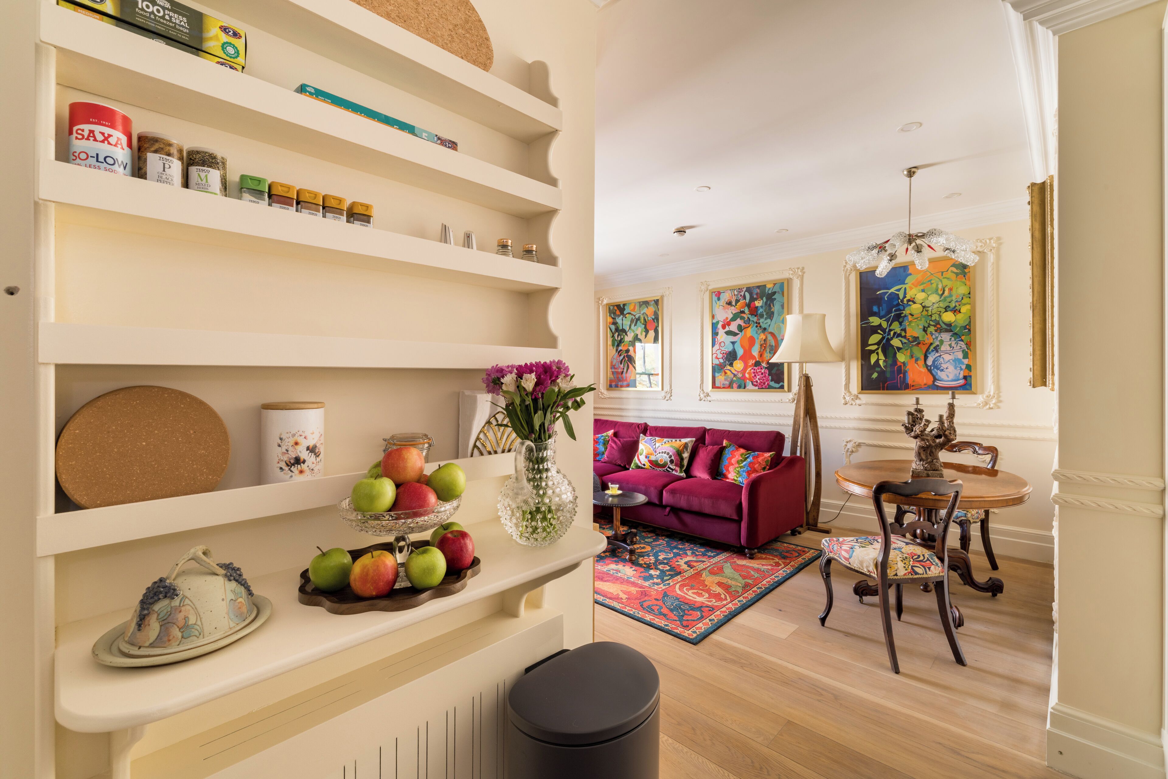 A living room with a sofa, a dining table with chairs, a vase with flowers, and shelves with various items.