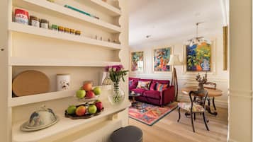 A living room with a sofa, a dining table with chairs, a vase with flowers, and shelves with various items.