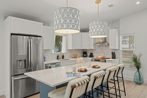 Una cocina moderna con un refrigerador de acero inoxidable, una isla de mármol, taburetes de bar, luces colgantes y un ventanal con persianas.
