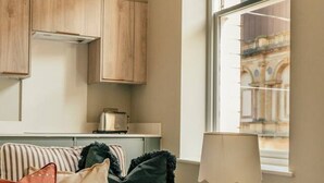Een gestreepte bank met kussens, een klein bijzettafeltje met een lamp, en een raam met uitzicht op een gebouw.