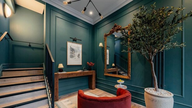 Een trap die leidt naar een kamer met teal muren, een houten console tafel, een rode poef, een grote spiegel en een potplant.