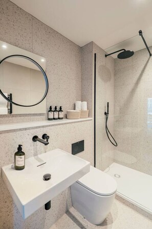 Une salle de bain moderne avec un lavabo, des toilettes et une douche, le tout de couleur blanche. Un miroir est placé au-dessus du lavabo, et une étagère contient des articles de toilette.