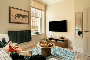 A living room with a sofa, a coffee table, a TV, a radiator, and a window with a view of buildings. - Stunningly Designed Central 1-Bed Apartment (North Yorkshire)