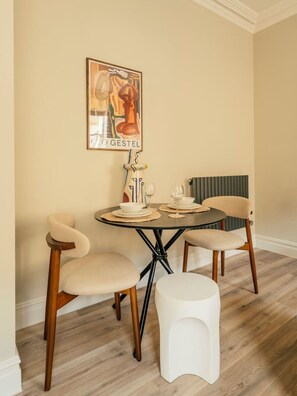 Un coin repas comprenant une table ronde, deux chaises, un tabouret blanc, une œuvre d’art encadrée et un radiateur.