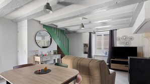 A modern living room with a wooden dining table, a sofa, a TV, and a staircase. - Gite de la Ferme de la Dalbeine – 25 min from Chenonceaux (Veuzain-sur-Loire)