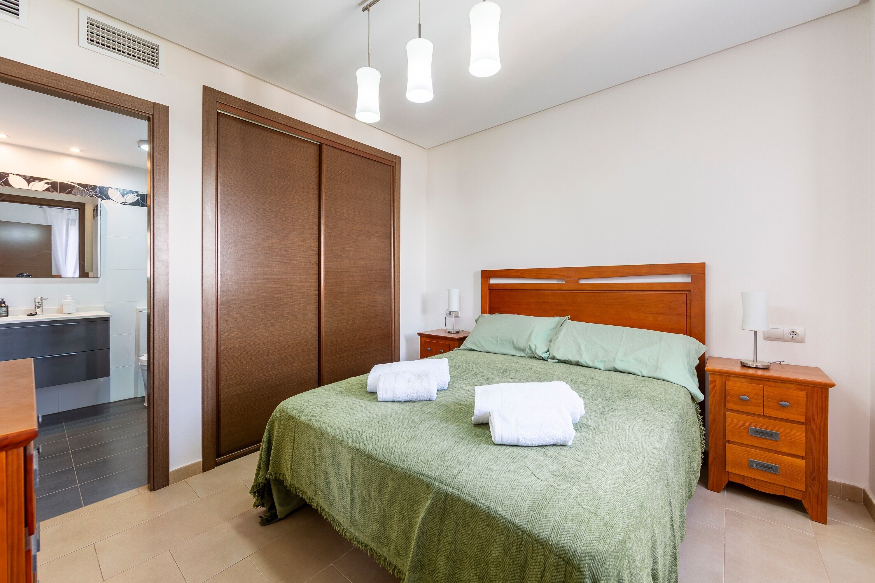 Un dormitorio con cama, mesitas de noche, un armario de madera y un baño a la vista a través de una puerta abierta.