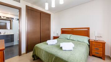 Un dormitorio con cama, mesitas de noche, un armario de madera y un baño a la vista a través de una puerta abierta.