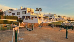 Zona de gimnasio al aire libre con equipo de ejercicio, un sendero pavimentado y un edificio con varios balcones y señalización al fondo.