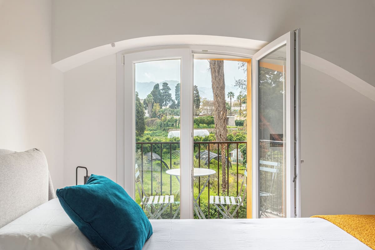Quarto com cama, travesseiro azul-esverdeado e varanda com mesa e cadeiras.