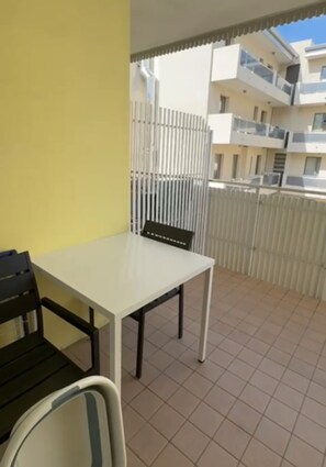 Un balcón con una mesa y sillas blancas, una pared amarilla y vistas a una cerca blanca y edificios.