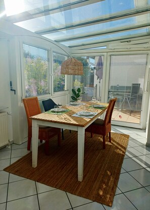 Un coin repas avec une table dressée pour quatre personnes, une chaise en osier, un fauteuil rembourré et un luminaire suspendu au plafond, fait de fibres tressées.