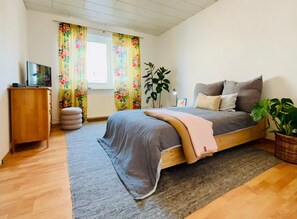 Une chambre à coucher avec un lit, une armoire en bois, un téléviseur, une fenêtre avec des rideaux à motifs floraux et une plante en pot.