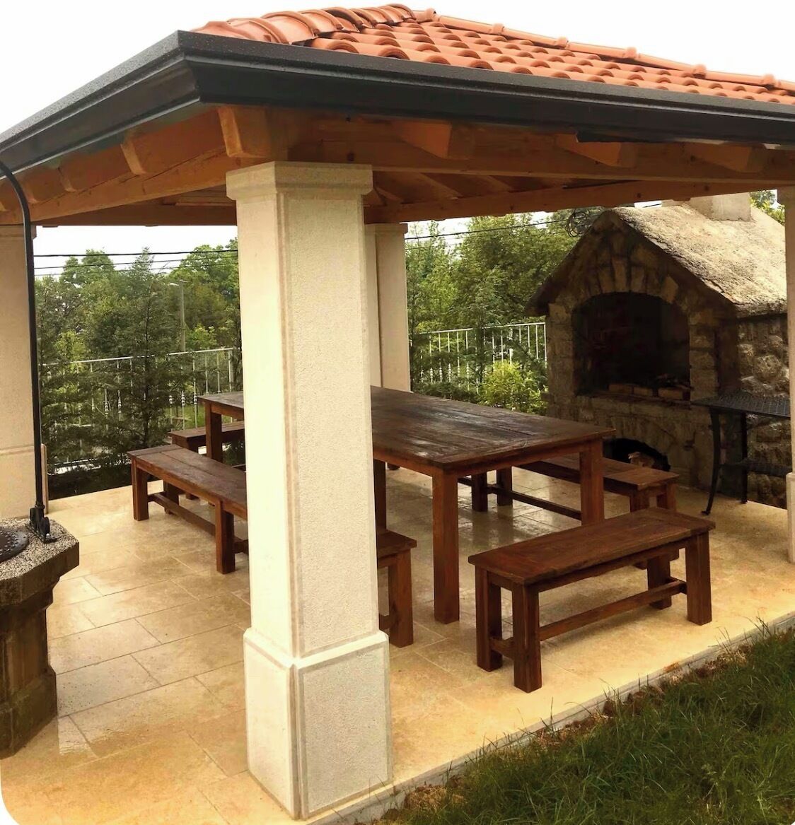 An outdoor patio with a stone fireplace, wooden tables and benches, and a view of trees.
