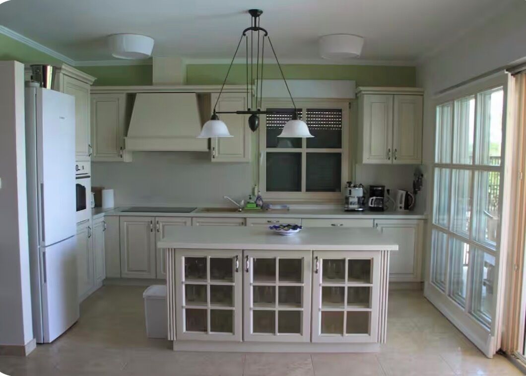 A kitchen with white cabinets, a central island with glass doors, pendant lighting, and a window on the right.