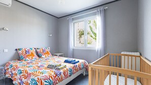 Un dormitorio con una cama, una cuna, una ventana con cortinas y una mesita de noche.
