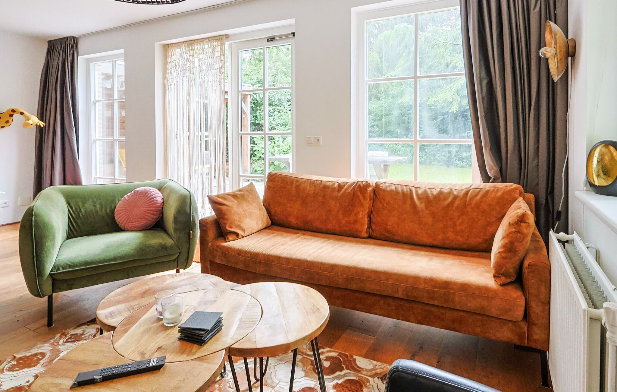 A living room with a green armchair, an orange sofa, a small round table with a remote control, and a window with curtains.