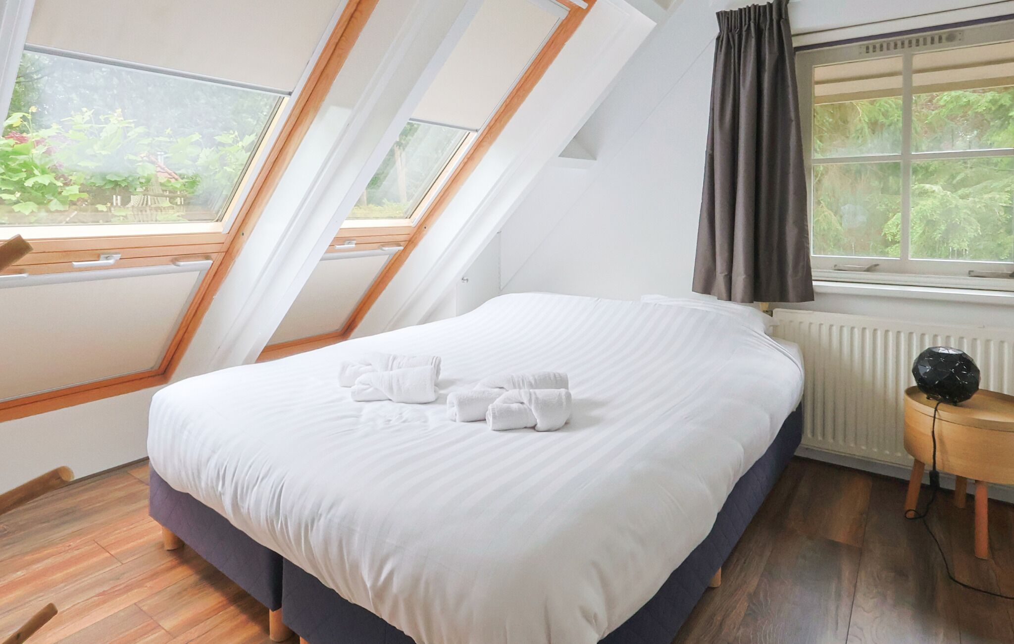 A bedroom with a bed, a window with curtains, a radiator, and a small table with a lamp.