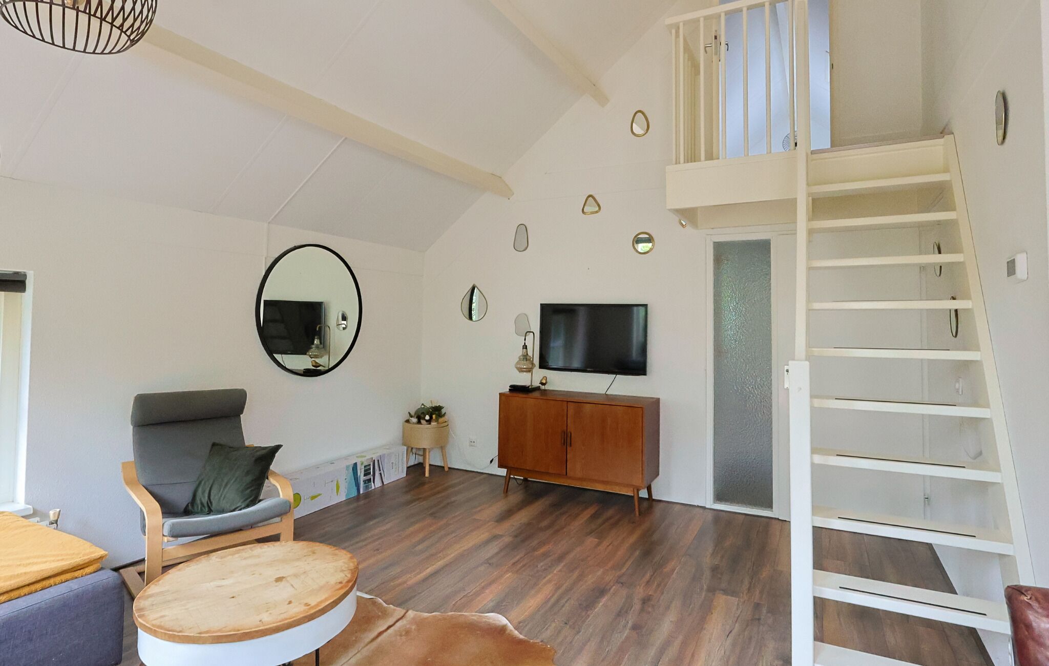 Amplia sala de estar con piso de madera, un sofá, una mesa de centro redonda, un televisor sobre un mueble de madera y una escalera blanca.